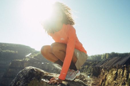 Frau mit lockigem Haar und einem orangen T-Shirt hockt auf einem Felsen vor einer bergigen Landschaft.